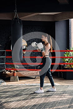 Young beautiful sports girl in sportswear and boxing gloves hits a punching bag in modern black gym.