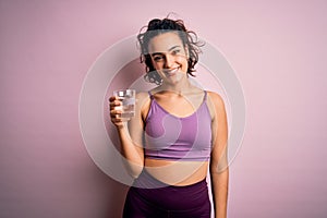 Young beautiful sportswoman with curly hair doing sport drinking glass of water with a happy face standing and smiling with a