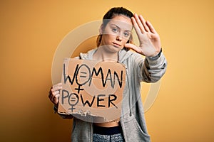 Young beautiful sportswoman holding woman power banner over isolated yellow background with open hand doing stop sign with serious