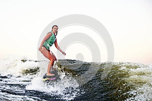 Young beautiful sporty girl in a green life jacket rides a wakeboard on a river or lake near city. Happy athlete girl