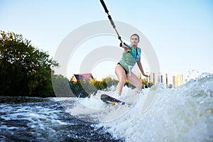 Young beautiful sporty girl in a green life jacket rides a wakeboard on a river or lake near city. Happy sportswoman