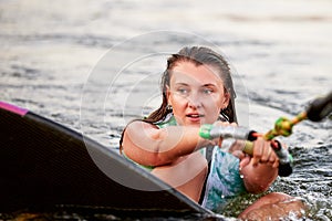 Young beautiful sporty girl in a green life jacket sitting in the water on a wakeboard with a halyard in her hand