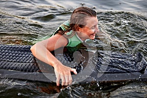 Young beautiful sporty girl in a green life jacket swims in the water with a wakeboard in her hand. Happy sportswoman is