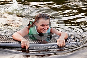 Young beautiful sporty girl in a green life jacket swims in the water with a wakeboard in her hands. Happy sportswoman