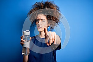 Young beautiful sporty woman with curly hair and piercing doing sport holding bottle of water pointing with finger to the camera