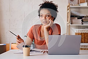 Young beautiful student girl with dark curly hair sitting at the table with laptop and cup of coffee to go leaning on