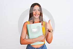 Young beautiful student girl holding notebook standing over isolated white background happy with big smile doing ok sign, thumb up