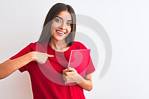 Young beautiful student woman holding red book standing over isolated white background with surprise face pointing finger to