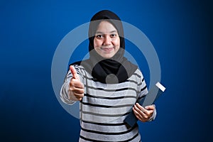 Young beautiful student woman wearing muslim hijab holding books doing happy thumbs up gesture with hand