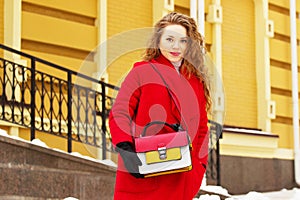 Young, beautiful and stylish blonde hair girl in red coat and with handbag walking through city streets. Womens fashion.