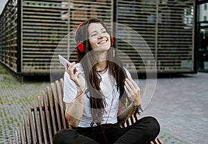 Young beautiful stylish brunette in white T-shirt uses her phone and headphones to listen to music online. Girl is sitting on