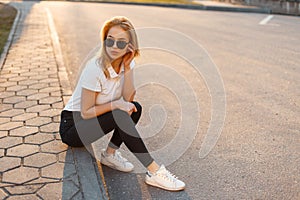 Young beautiful stylish hipster woman with sunglasses in a white polo shirt and black jeans with sneakers sitting on the street at