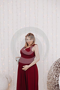 Young beautiful stylish pregnant woman in red long evening dress on white background of child room