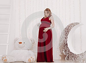 Young beautiful stylish pregnant woman in red long evening dress on white background of child room