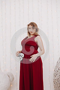 Young beautiful stylish pregnant woman in red long evening dress on white background of child room