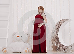 Young beautiful stylish pregnant woman in red long evening dress on white background of child room