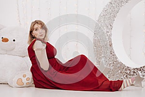 Young beautiful stylish pregnant woman in red long evening dress on white background of child room