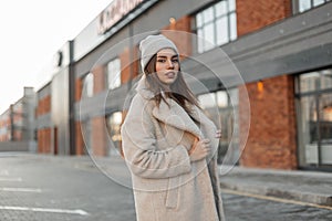 Young beautiful stylish woman in a fashionable knitted knitted hat in a trendy eco fur coat poses in a parking lot near a vintage