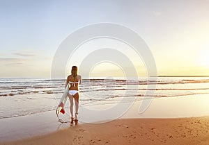 Young beautiful surfer girl walking towards surf at sunrise