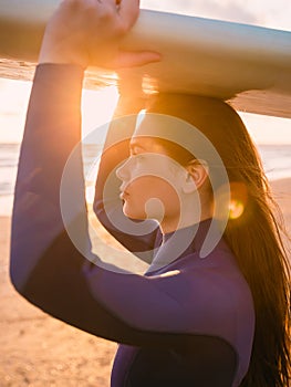 Young beautiful surfgirl with long hairs and in wetsuit. Posing with surf board near ocean at sunset. Ready for surfing.