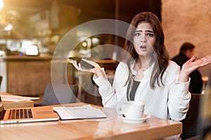 Young beautiful surprised woman with phone in her hand working outside office in laptop looks desperate drinking hot coffee in