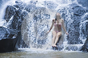 Young beautiful and sweet Asian woman in bikini getting body wet under stream of natural amazing waterfall sitting on rock feeling