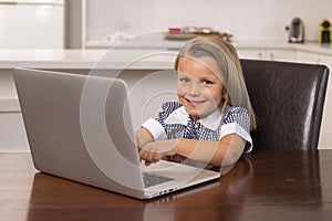 Young beautiful and sweet little girl 6 to 8 years old with blond hair and blue eyes sitting at home kitchen enjoying with laptop
