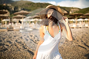 Young beautiful tanned brunette woman wearing hat and elegant dress standing on beach with wind fluttering hair.
