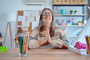 Young beautiful teacher woman wearing sweater and glasses sitting on desk at kindergarten begging and praying with hands together