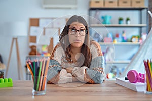 Young beautiful teacher woman wearing sweater and glasses sitting on desk at kindergarten looking sleepy and tired, exhausted for
