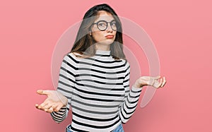 Young beautiful teen girl wearing casual clothes and glasses clueless and confused expression with arms and hands raised