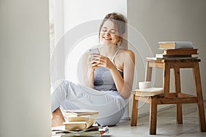 Young beautiful teenage girl surfing web looking at phone screen smiling sitting on floor among old books near window