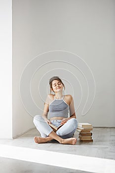 Young beautiful tender girl smiling with closed eyes holding book sitting on floor over white wall early in morning.