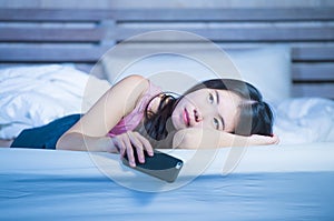 Young beautiful and thoughtful Asian Chinese woman on her 20s holding mobile phone lying on bed at night thinking looking sad and