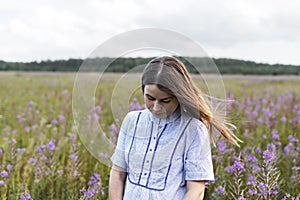 Young beautiful thoughtful blond woman in purple shirt walking in the meadow among flowers of fireweed