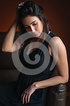 Young beautiful thoughtful woman titivating long dark hair in black dress sit reclining on brown sofa with closed eyes.