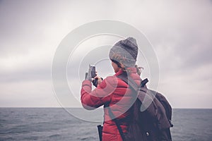 Young beautiful traveler girl with a backpack taking off the ocean and waves on a smartphone, winter, Atlantic ocean, Portugal