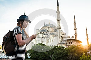 A young beautiful traveler girl in a hat with a backpack is watching a map next to the blue mosque - the famous sight of