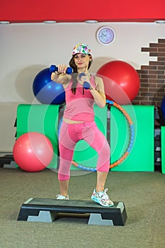 Young beautiful white girl in a pink sports suit does physical exercises with dumbbells at the fitness center.