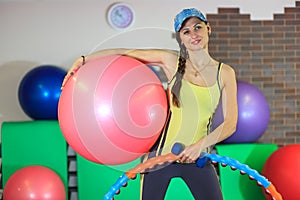 Young beautiful white girl in a yellow and gray sports suit stands with a fit ball, hoop and a dumbbell at the fitness center.