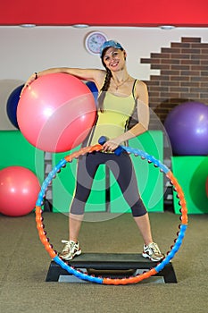 Young beautiful white girl in a yellow and gray sports suit stands with a fit ball, hoop and a dumbbell at the fitness center.