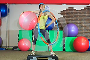 Young beautiful white girl in a yellow and gray sports suit stands with a fit ball, hoop and a dumbbell at the fitness center.