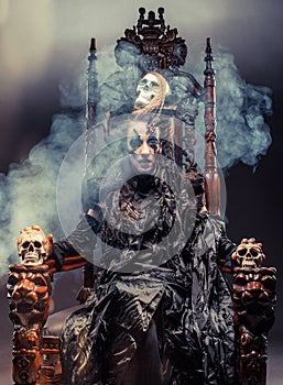 Young beautiful witch sits on a chair. Bright make up, skull, smoke- halloween theme.
