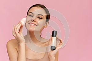 Young beautiful woman applying foundation with sponge and looking at camera on pink background. Perfect skin, makeup