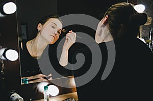 Young beautiful woman applying her make up face with brush, looking in a mirror, sitting on chair at dressing room with