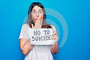 Young beautiful woman asking for psychical problem holding paper with not to suicide message covering mouth with hand, shocked and