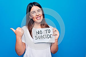 Young beautiful woman asking for psychical problem holding paper with not to suicide message pointing thumb up to the side smiling