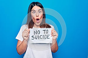 Young beautiful woman asking for psychical problem holding paper with not to suicide message scared and amazed with open mouth for