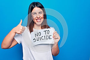 Young beautiful woman asking for psychical problem holding paper with not to suicide message smiling happy and positive, thumb up