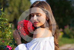 Young beautiful woman with bare shoulders in white in the garden with a red roses